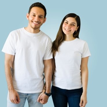 A man and woman wearing matching white t-shirts stand together, smiling and holding hands against a light blue background.