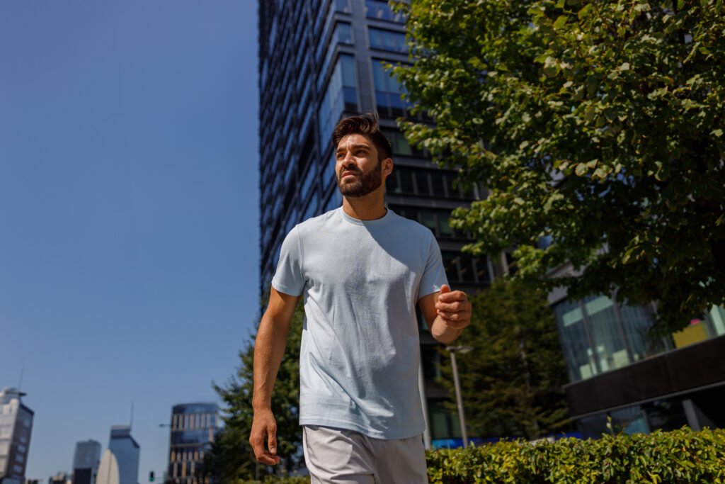 A man walking outdoors in a light blue t-shirt and shorts, with modern buildings and trees in the background under a clear blue sky.