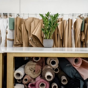 A workspace with a variety of rayon fabric rolls stored underneath a table, while patterns hang on a rack in the background. A small green plant is placed in a white pot on the table. "does rayon fabric shrink"