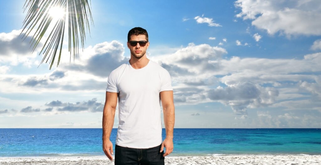 A man wearing a plain white t-shirt and stands on a beach with a beautiful ocean view and palm tree in the foreground.