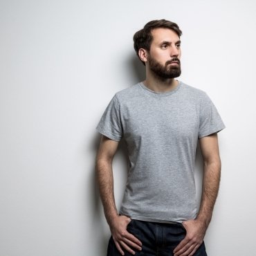 A man wearing a gray t-shirt stands against a plain white wall, with a thoughtful expression and hands on his hips.