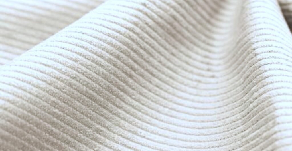 Close-up view of a textured corduroy fabric in a light color, showing its distinctive ribbed pattern.