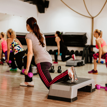A group of women in colorful trendy workout outfits participating in a fitness class. One woman is performing a lunge with dumbbells while others are engaged in different exercises.