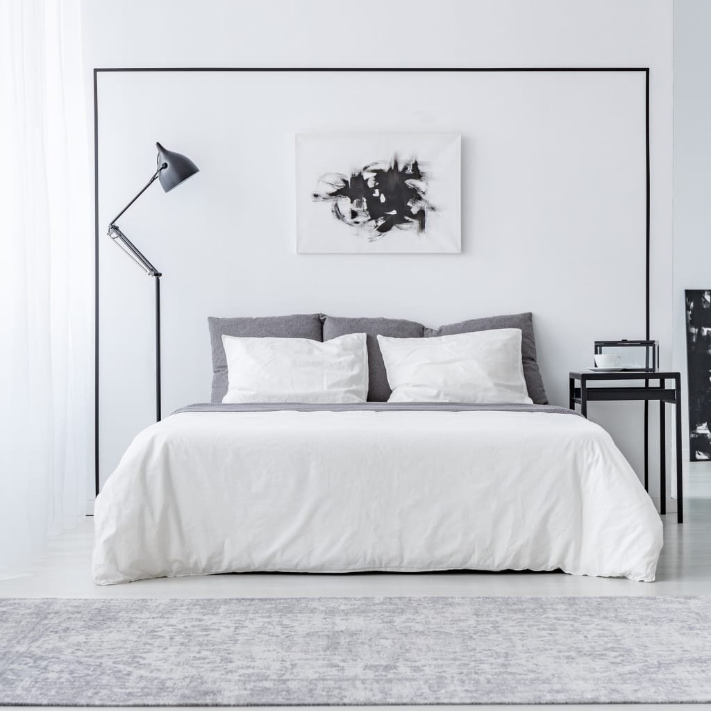  A minimalist bedroom with a white bed featuring gray and white pillows, a black framed artwork above, and a side table with a small decoration. A black adjustable floor lamp stands next to the bed, and the room has a clean, modern aesthetic with mostly white and black tones.