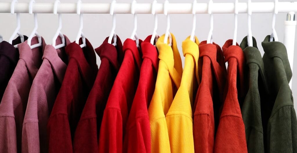 A row of colorful hoodies hanging on a white clothing rack, arranged in a gradient from dark purple to green.