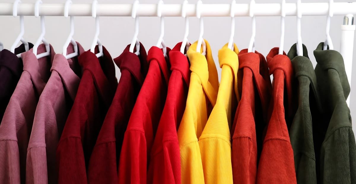 A row of colorful hoodies hanging on a white clothing rack, arranged in a gradient from dark purple to green.