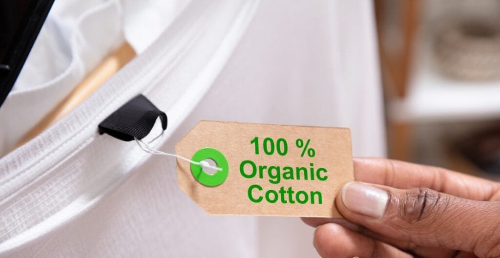 A hand holding a brown tag that reads "100% Organic Cotton" attached to a white fabric product with a black anchoring loop