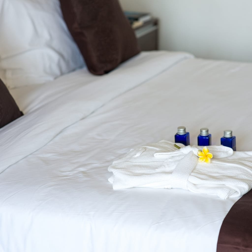A neatly made bed with white bedding, dark brown pillows, and a stack of white towels adorned with a yellow flower, alongside three small blue bottles placed on the bed.