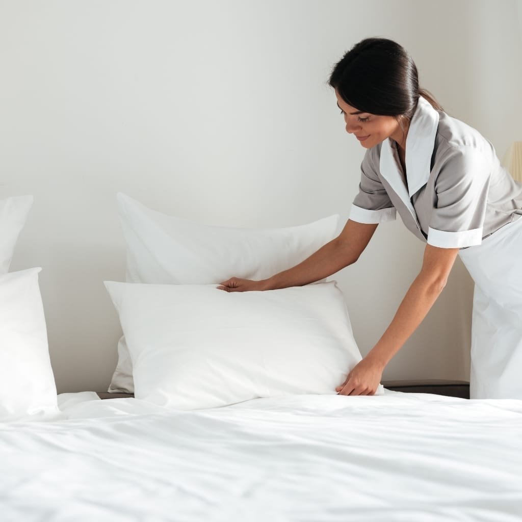 A woman dressed in a gray uniform, likely a housekeeper or cleaner, is making a white bed with neatly arranged pillows in a bright, minimal bedroom.