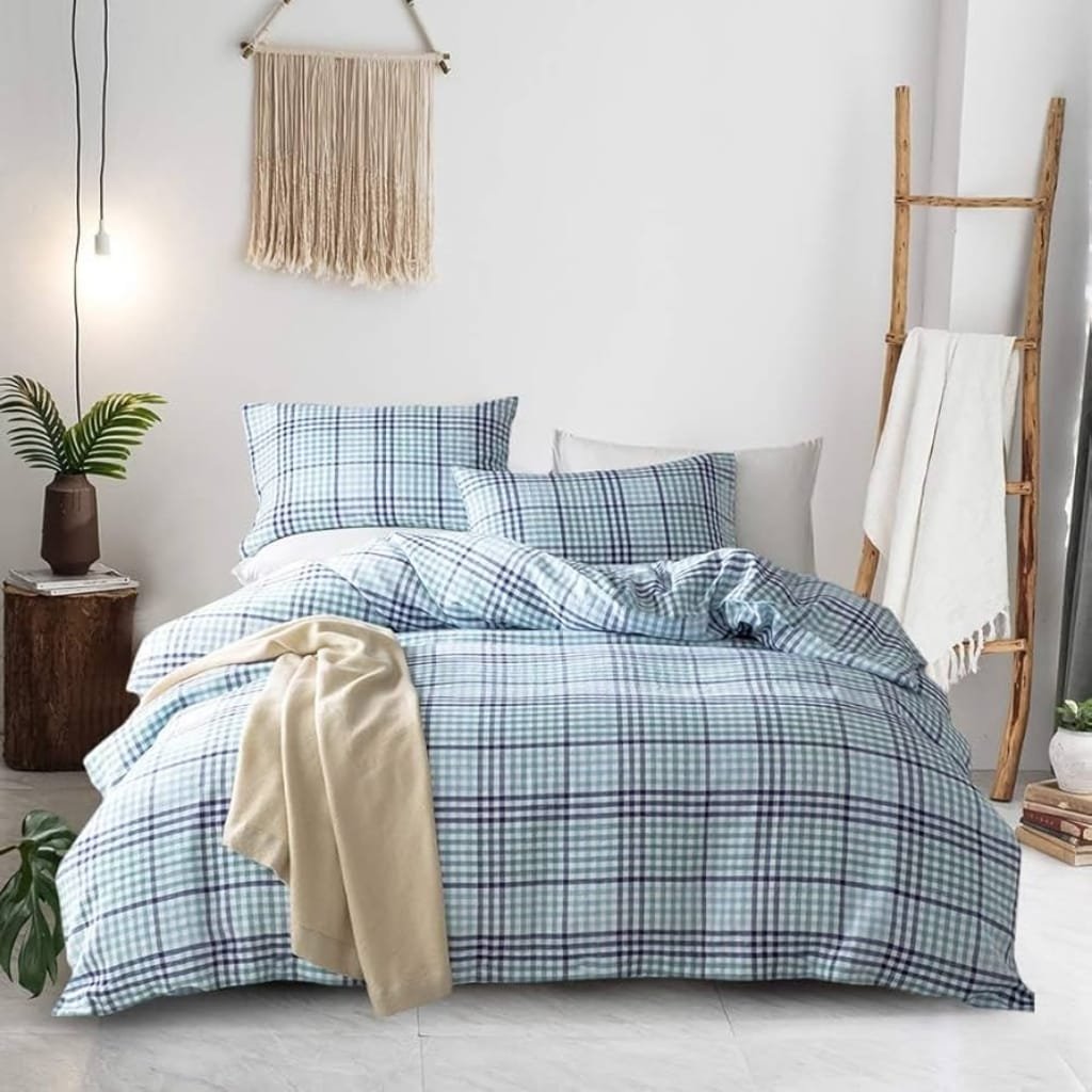 A cozy bedroom with a bed covered in plaid bedding, featuring multiple pillows and a beige throw blanket. There is a wooden side table with a plant, a macrame wall hanging, a wooden ladder with a white towel, and a window with natural light filtering in.