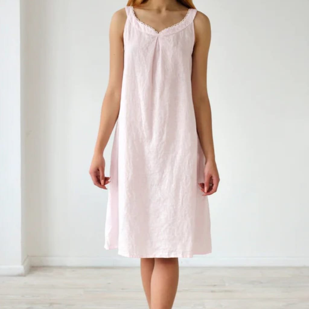 A woman wearing a light pink, sleeveless, knee-length nightgown with a V-neckline and lace trim, standing against a plain white wall.