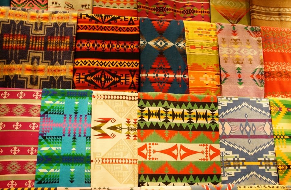 A colorful collection of various woven textiles with geometric patterns, including stripes, diamonds, and intricate motifs in vibrant shades of red, yellow, blue, green, orange, and beige.