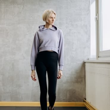 A woman standing indoors next to a window, wearing black polyester leggings with a matching hoodie, showcasing a comfortable athleisure outfit.