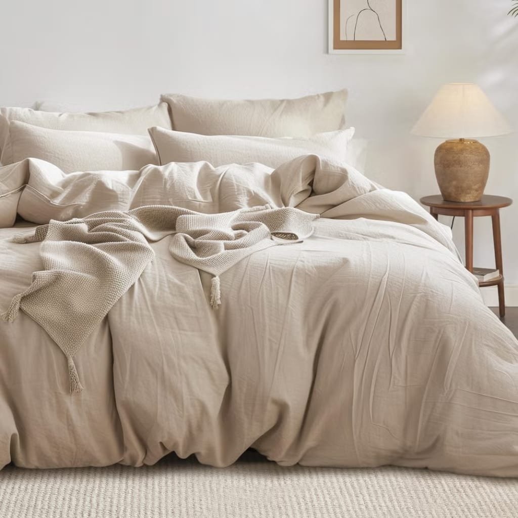 A cozy, neatly made bed with off-white, textured bedding and multiple pillows. A soft blanket is casually draped over the bed. To the right, there is a wooden side table with a large, earthy-toned lamp, and a simple, minimalistic framed line drawing on the wall behind.