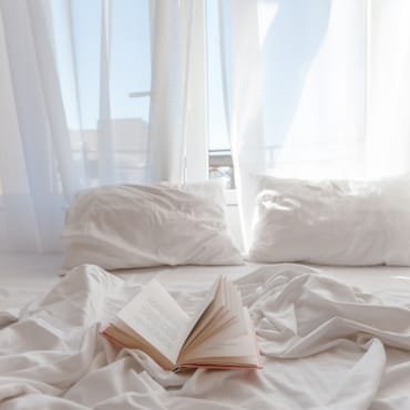 Sunlit bedroom scene with a white bed, pillows, and an open book, showcasing soft bedding.