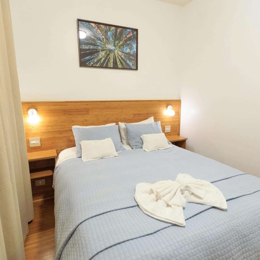 A neatly made double bed in a cozy hotel room with a wooden headboard, two bedside tables with wall-mounted lamps, a framed picture of trees on the wall above, and a towel folded into a decorative shape on the bed.