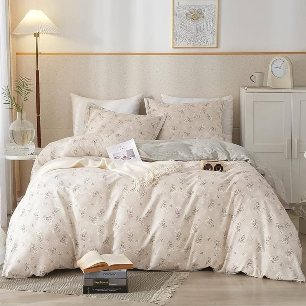 A cozy, neatly made bed with cream-colored floral-patterned bedding, topped with several pillows. To the right of the bed is a white cabinet with a mug, clock, and books. A small table with a glass vase and a book sits to the left. A book and a box of espresso capsules are on the floor in front of the bed. The room has soft lighting from a standing lamp and simple decor, including a framed picture above the bed.