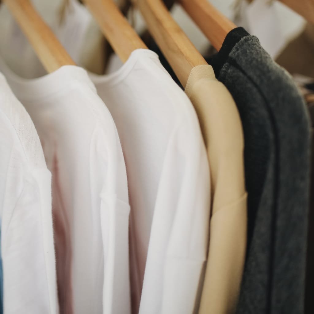 Close-up of hanging garments made of cotton, including white, beige, and dark gray shirts on wooden hangers.