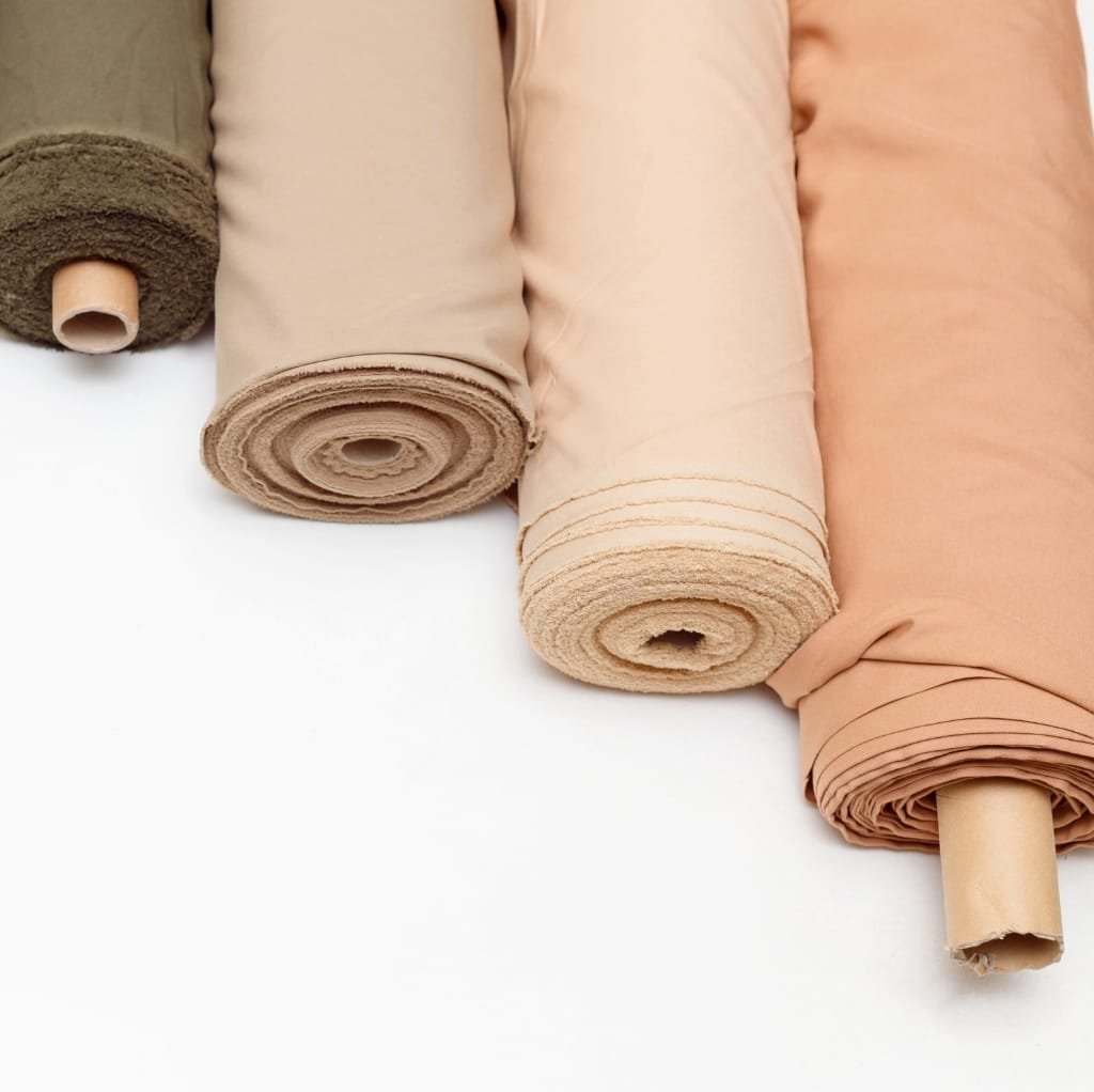 Four rolls of cotton fabric in shades of green, beige, light peach, and tan, arranged side by side on a white background.