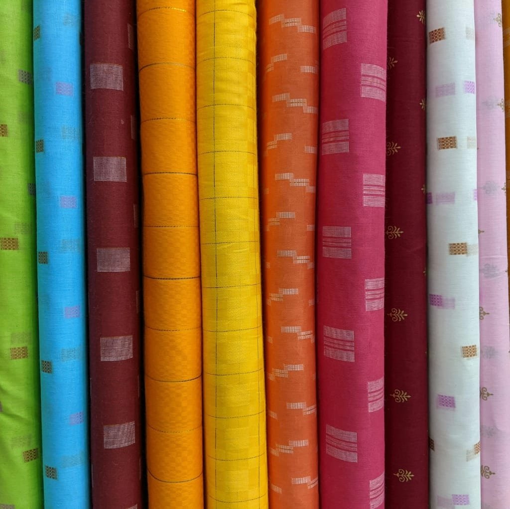 Colorful rolls of poplin cotton fabric aligned vertically, showcasing shades of green, blue, maroon, yellow, orange, pink, and white with various subtle patterns.