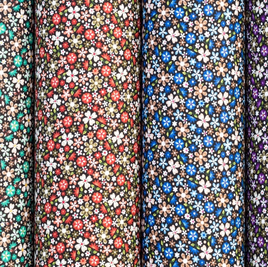 Close-up image of multiple rolls of cotton fabric with floral patterns, arranged vertically. The fabrics feature various colorful flower designs on dark backgrounds.