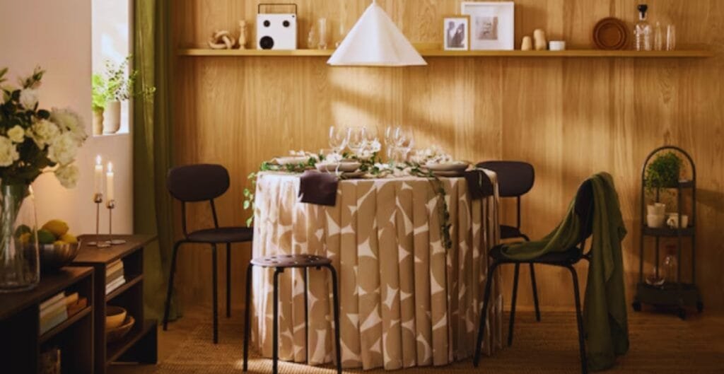 A cozy dining area featuring a round table draped with a beige and white patterned tablecloth, surrounded by four black chairs. The table is set with glassware and cutlery, and there are candles on a small side table to the left. The background has wooden paneled walls with a floating shelf holding framed photos and decorative items. Soft light fills the room, creating a warm atmosphere.