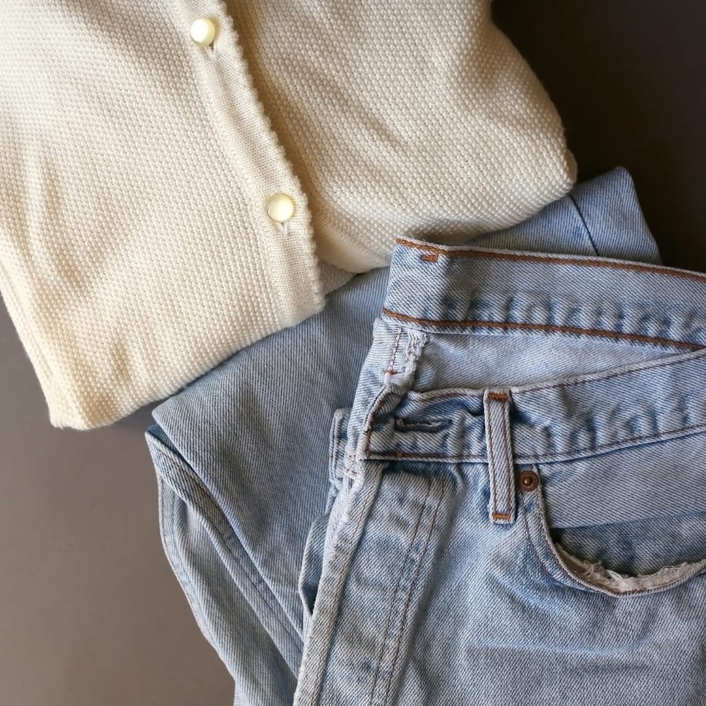 A close-up of a light-colored textured cardigan with pearl-like buttons, partly revealing a pair of light blue, slightly distressed jeans with fraying near the pocket and waistband.