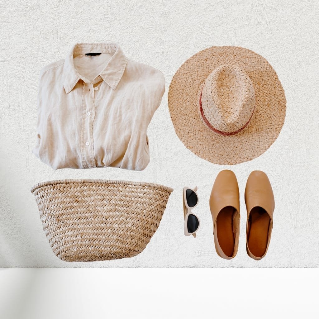 A flat lay of summer accessories and clothing items on a white textured surface, including a beige button-up shirt, a wide-brimmed straw hat, a woven straw bag, a pair of tan slip-on shoes, and a pair of white sunglasses with black lenses.