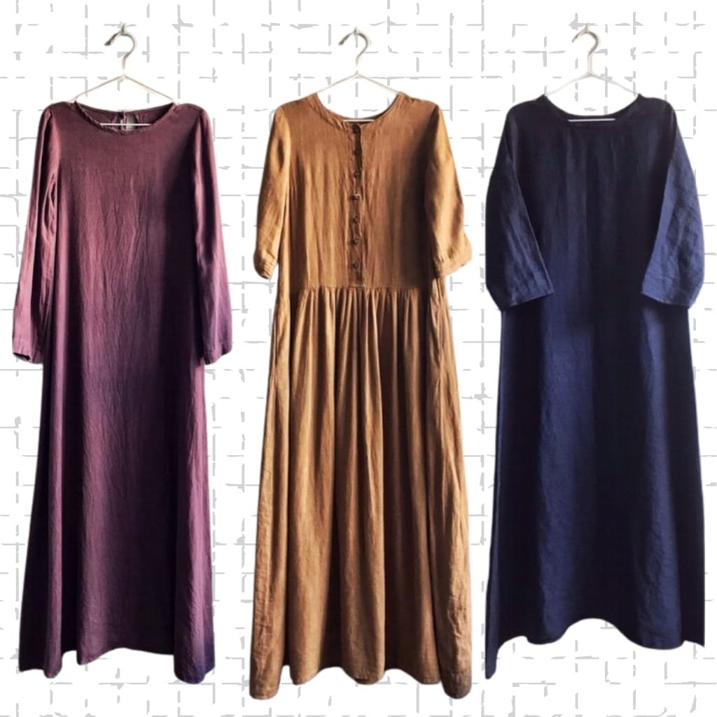 Three long dresses hanging on wall-mounted hangers against a white brick background. The first dress on the left is a long-sleeved, burgundy gown. The middle dress is a mustard-colored, button-down, long-sleeved dress with a gathered skirt. The third dress on the right is a dark navy, long-sleeved dress with a simple, loose fit.