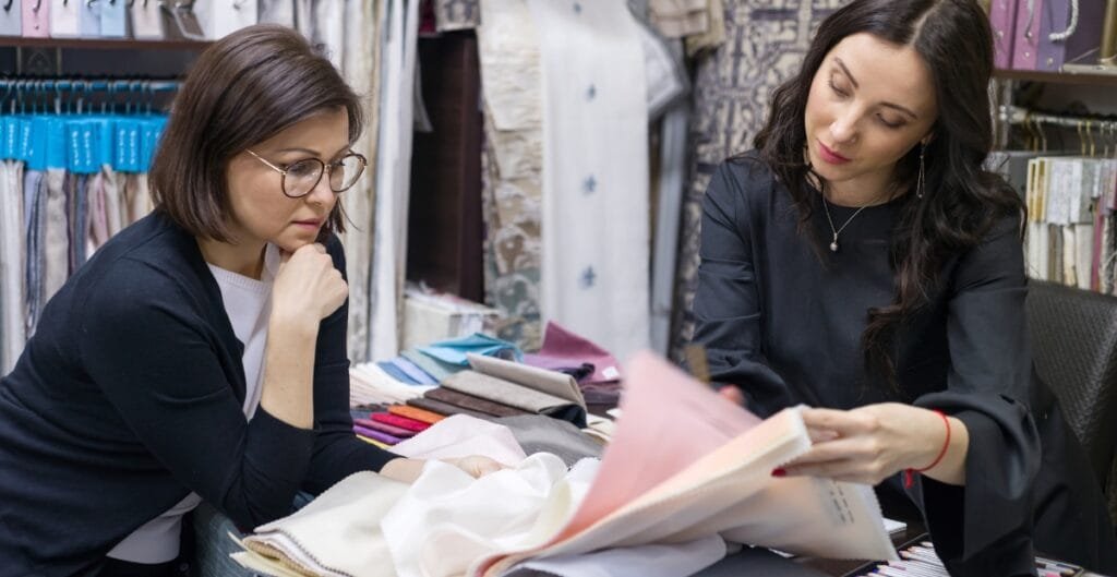 Two women are sitting at a table in a fabric or textile store, examining fabric samples and swatches carefully.
