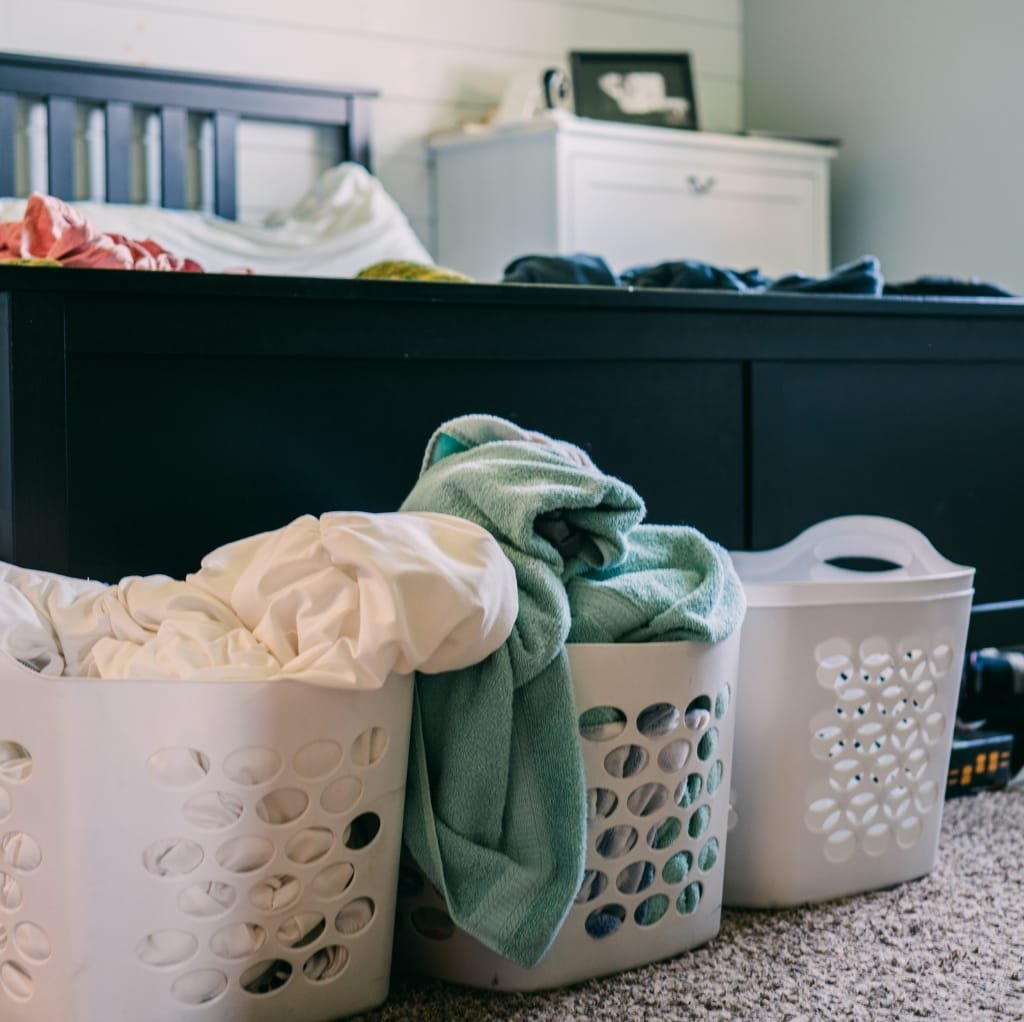 Three white laundry baskets filled with clothes, placed in front of a black bed frame and a white dresser in a bedroom.
