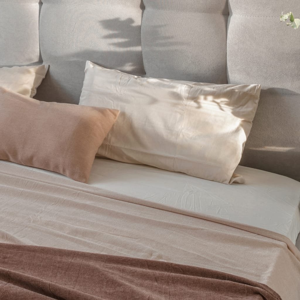 A bedroom scene with a soft, beige bedspread, large satin pillows, and a textured gray upholstered headboard. A branch with flowers casts a shadow on the pillows.