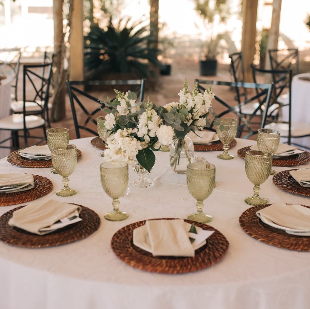A round table set for an elegant event, featuring a white tablecloth, woven placemats, neatly folded cream napkins, vintage-style green glass goblets, and a central floral centerpiece of white flowers and greenery in a clear glass vase. The background reveals a charming outdoor setting with natural light and wooden chairs. Bulk Cotton Fabric