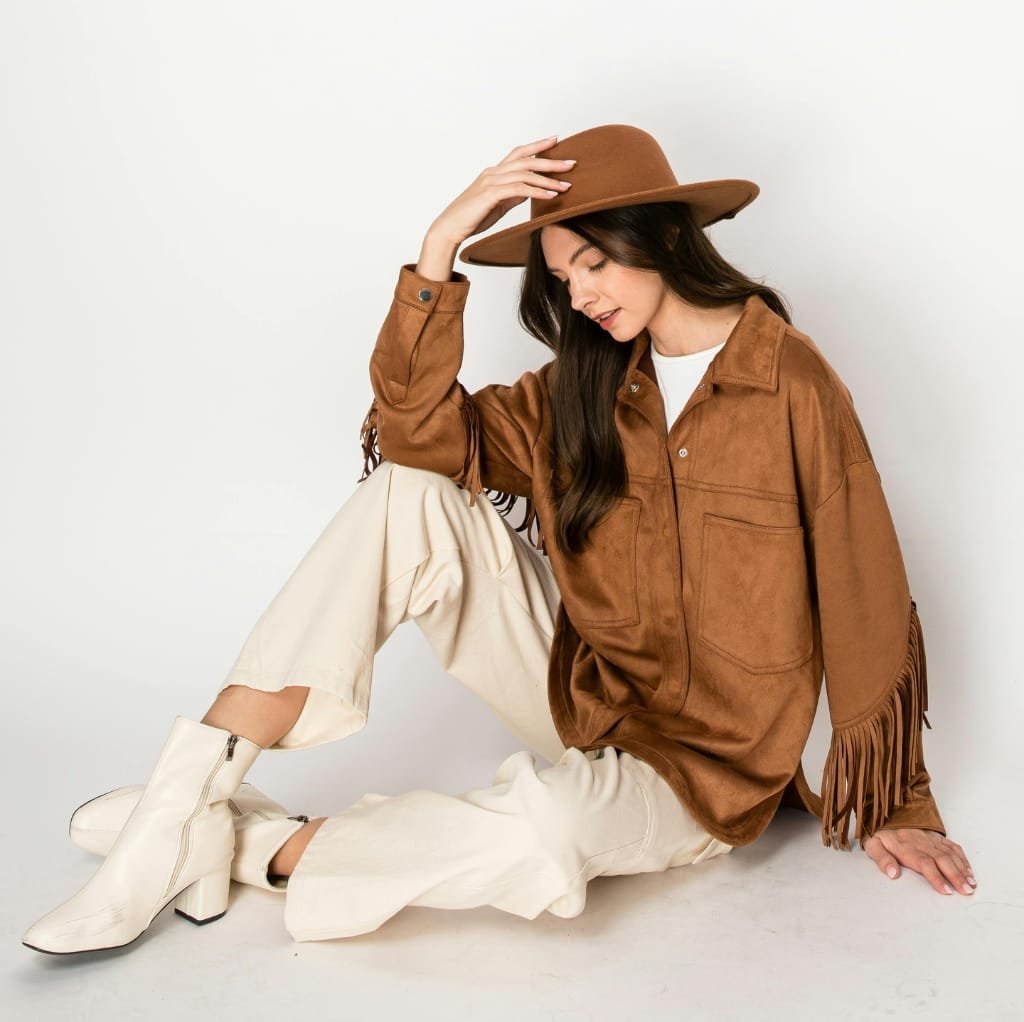 A young woman sitting on the floor against a plain white background, dressed in a stylish fall-inspired outfit with a brown hat, brown fringed jacket, cream-colored pants, and white ankle boots, gently touching her hat while looking down.