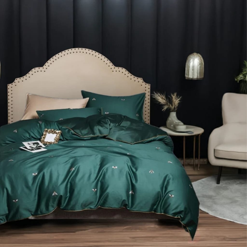 A stylish bedroom corner featuring a bed with dark green satin bedding and matching pillows, a cream-colored headboard with nailhead trim, a small round side table with a vase of dried flowers, a modern wall-mounted lamp, and a white armchair with a rounded back next to a light-colored rug on a wooden floor.