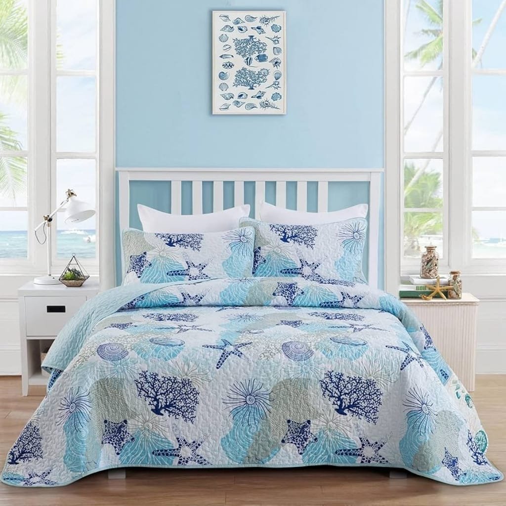 A cozy bedroom with a light blue wall and large windows showing a view of the ocean and palm trees outside. The bed has a white wooden headboard and is covered with a quilt and pillows featuring a marine-inspired pattern of starfish, sea shells, and coral in various shades of blue, teal, and cream. On each side of the bed are white nightstands, one with a modern white lamp and the other with small decorative jars, books, and sea-themed decor. A framed art print of various fish hangs on the wall above the bed.