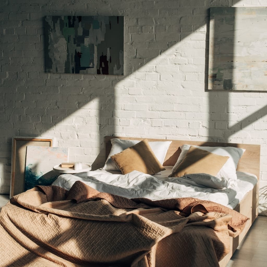 A modern bedroom with sunlight streaming through windows, casting shadows on a white brick wall. The bed has a light wooden headboard, with beige and gray pillows on a rumpled white sheet, and a brown quilted blanket draped at the foot of the bed. Several framed artworks rest against the wall beside the bed.