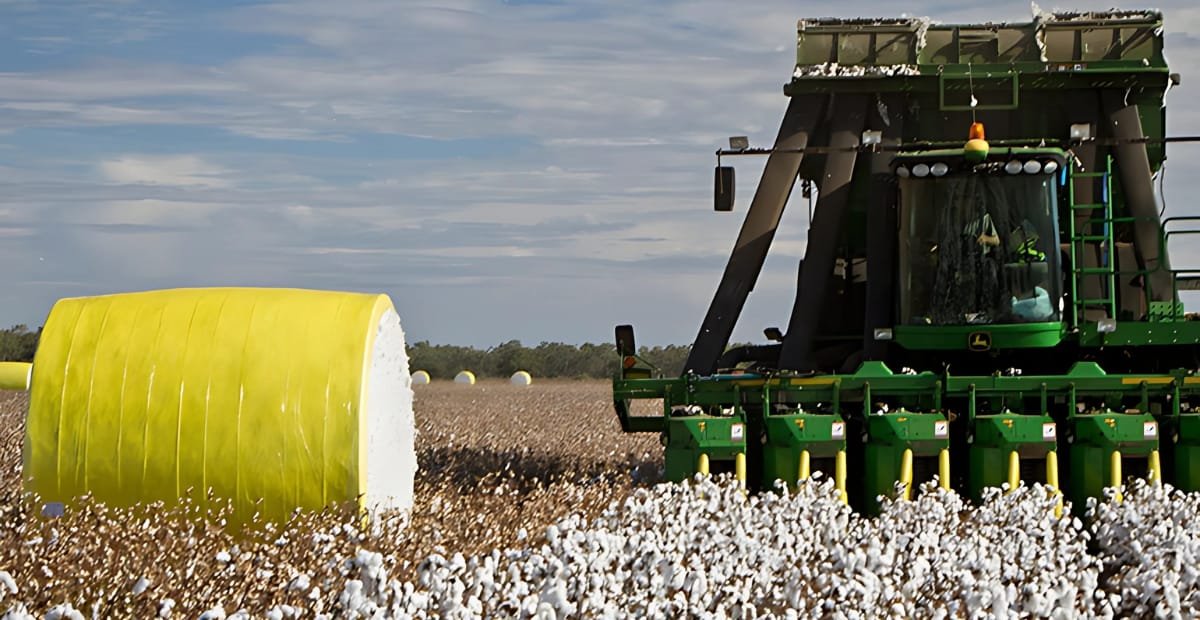 A green John Deere cotton picker machine harvesting cotton in a field with large yellow and white cotton bales in the background under a partly cloudy sky.