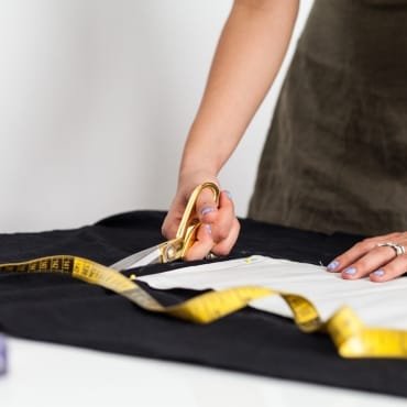 A person using scissors to cut a piece of black polyester fabric on a table, with a yellow measuring tape nearby.
