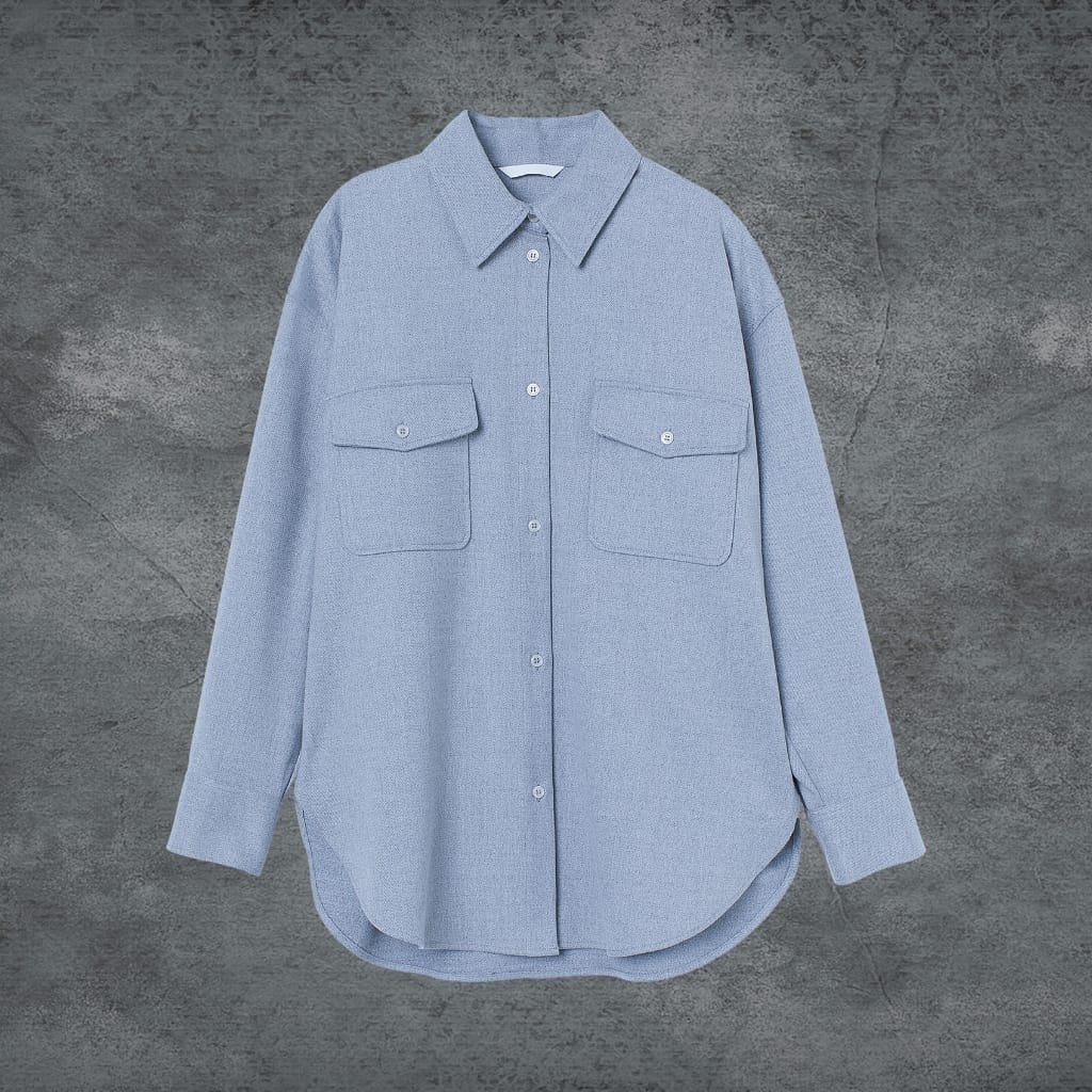 A light blue, long-sleeved button-up shirt with two chest pockets, laid flat against a textured gray background.