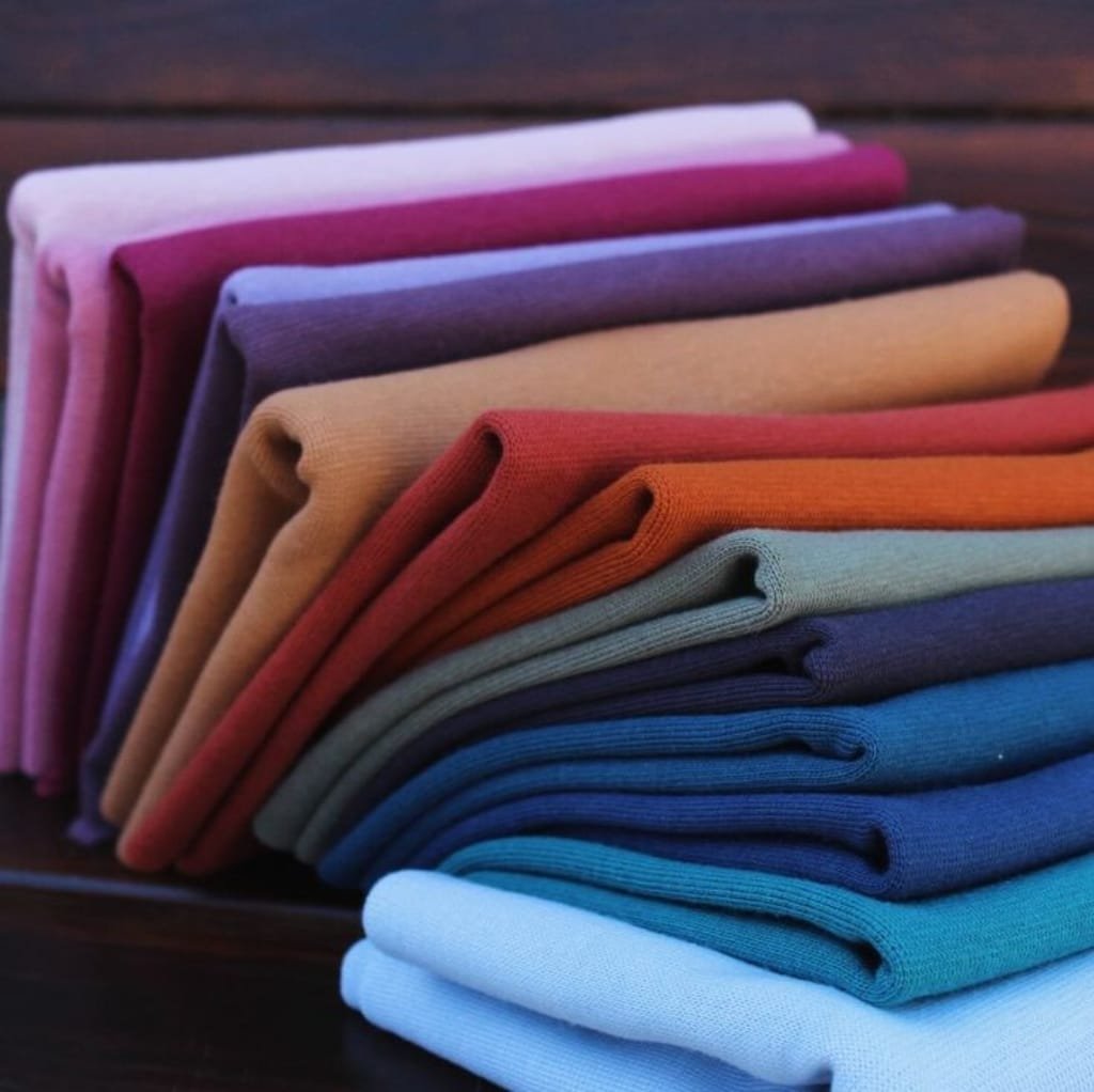 A colorful stack of folded jersey cotton fabric dresses in various shades, arranged on a dark wooden surface.