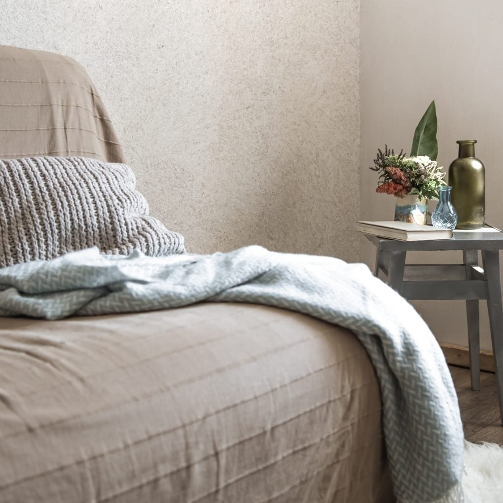 A cozy bedroom corner featuring a beige bed with a textured blanket and a chunky knit pillow, next to a small gray side table holding decorative vases, a book, and a flower arrangement.