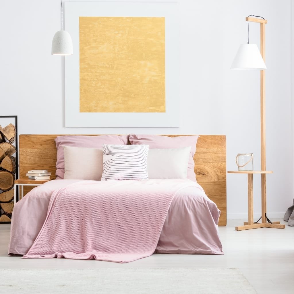 A cozy bedroom with a pink bedspread, multiple pillows, and a wooden headboard. There is a modern wooden standing lamp with a white lampshade to the right, a painting with a yellow square above the bed, and a stack of firewood on the left.