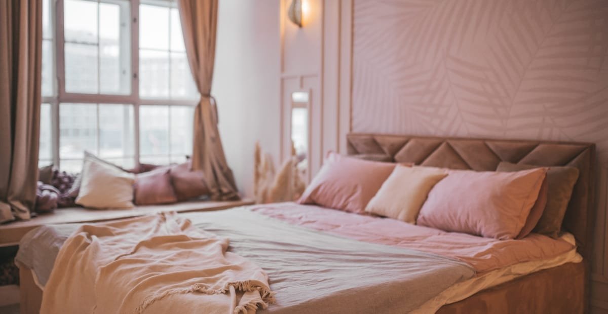 Cozy bedroom with a large bed, pink and cream pillows, and soft curtains over a window, illuminated by natural light.