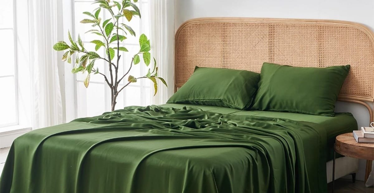 A neatly made bed with green satin sheets and matching pillows, set against a rattan headboard. To the left, a large indoor plant sits near a window with sheer white curtains, allowing natural light to fill the room. On the right, a small wooden bedside table holds a white telephone and a few books.