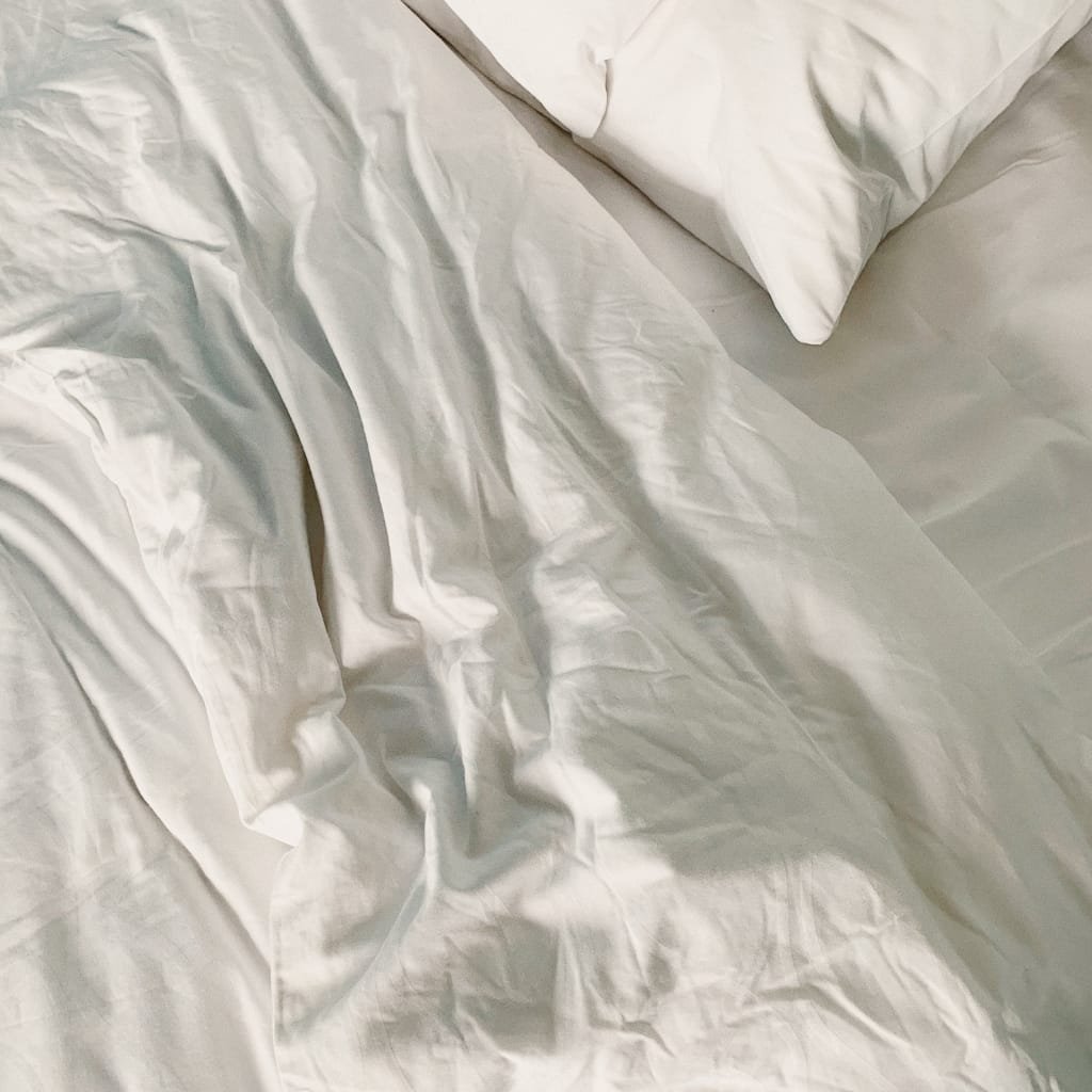 A close-up of wrinkled, cream-colored satin fabric on a bed, with a pillow partially visible in the top right corner.