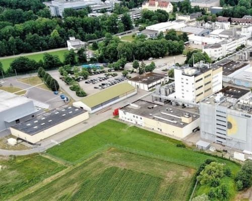 Aerial view of an industrial complex with surrounding green spaces and fields, featuring several buildings, parking areas, and covered storage facilities.