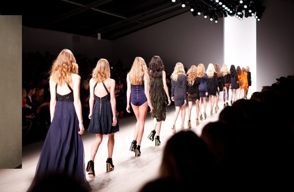 A line of models walking down the runway during a luxury fashion show, showcasing elegant designer outfits and high heels, with an audience watching in the background.