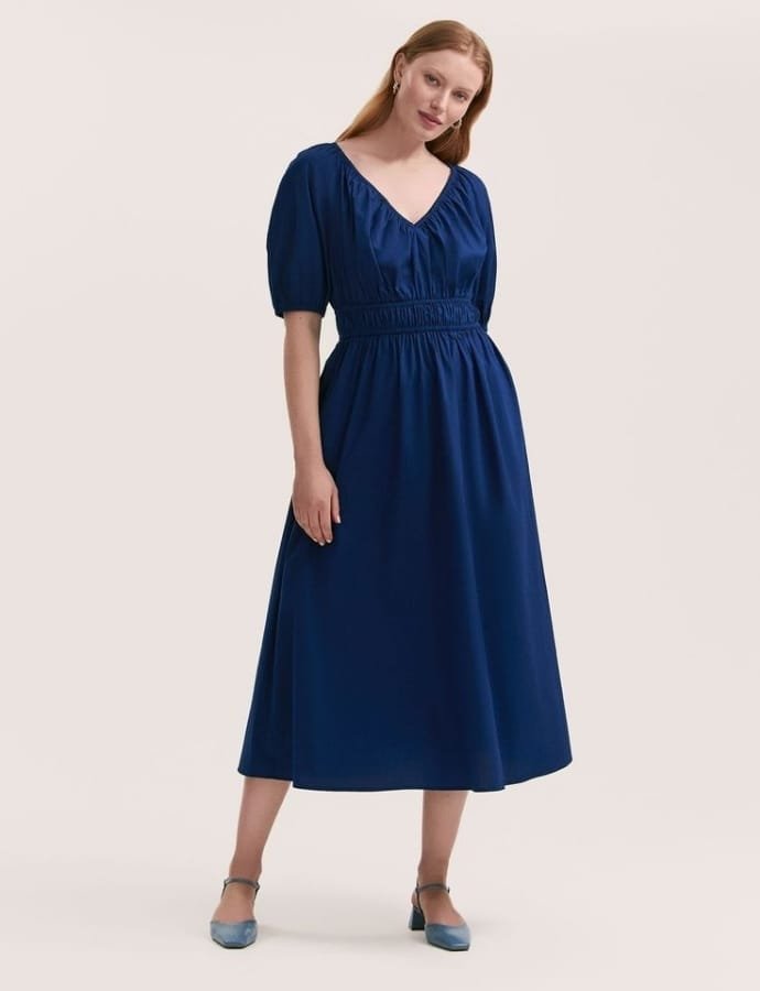 A woman wearing a cobalt blue midi cotton dress with puffed sleeves, a V-neckline, and an elastic waistband, paired with matching blue shoes.
