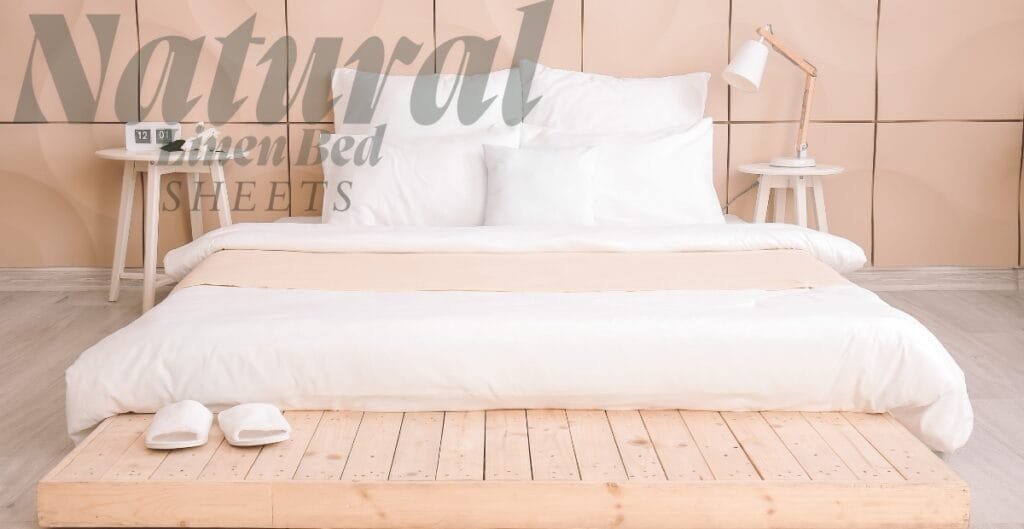A neatly made bed with white linens and pillows, set against a beige paneled wall. Two matching white bedside tables with lamps are on either side of the bed. There is a light wooden pallet at the foot of the bed with a pair of white slippers placed on it. Overlaid text reads "Natural Linen Bed SHEETS."